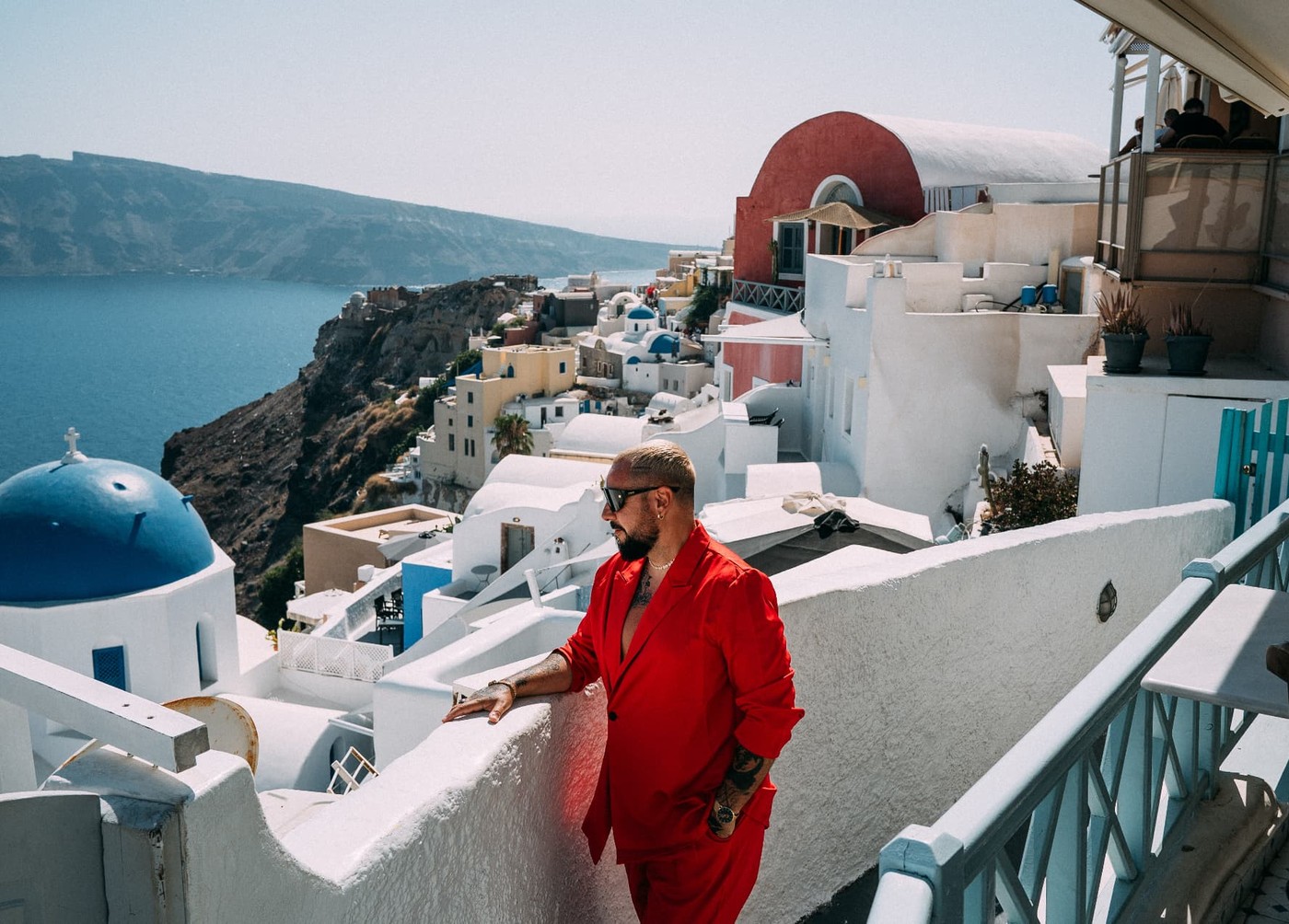 Marc Isla en costume rouge et lunettes de soleil, adossé à un muret blanc à Santorin, la mer et le village en arrière-plan