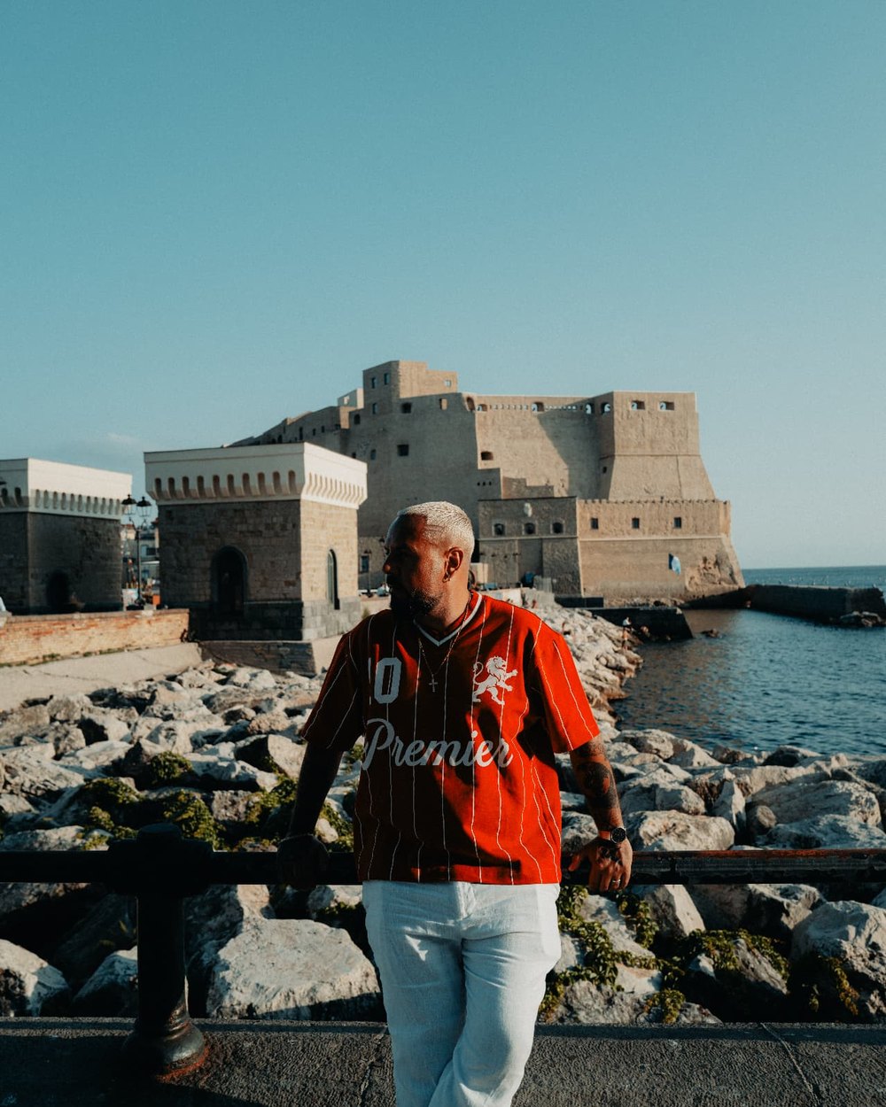 Marc Isla en veste rouge et lunettes noires, au bord de la mer, un château de pierre derrière lui